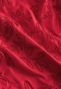 Tissu rouge avec une broderie florale complexe dans différentes nuances de rouge. Texture lisse avec des motifs de points en relief détaillés.