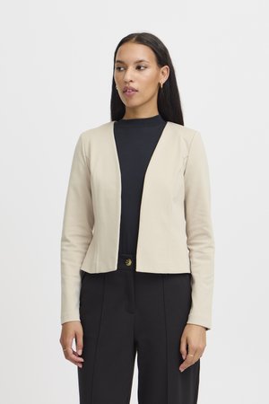Kvinde iført en beige åben front blazer over en sort top og sorte bukser, stående mod en ensfarvet hvid baggrund.