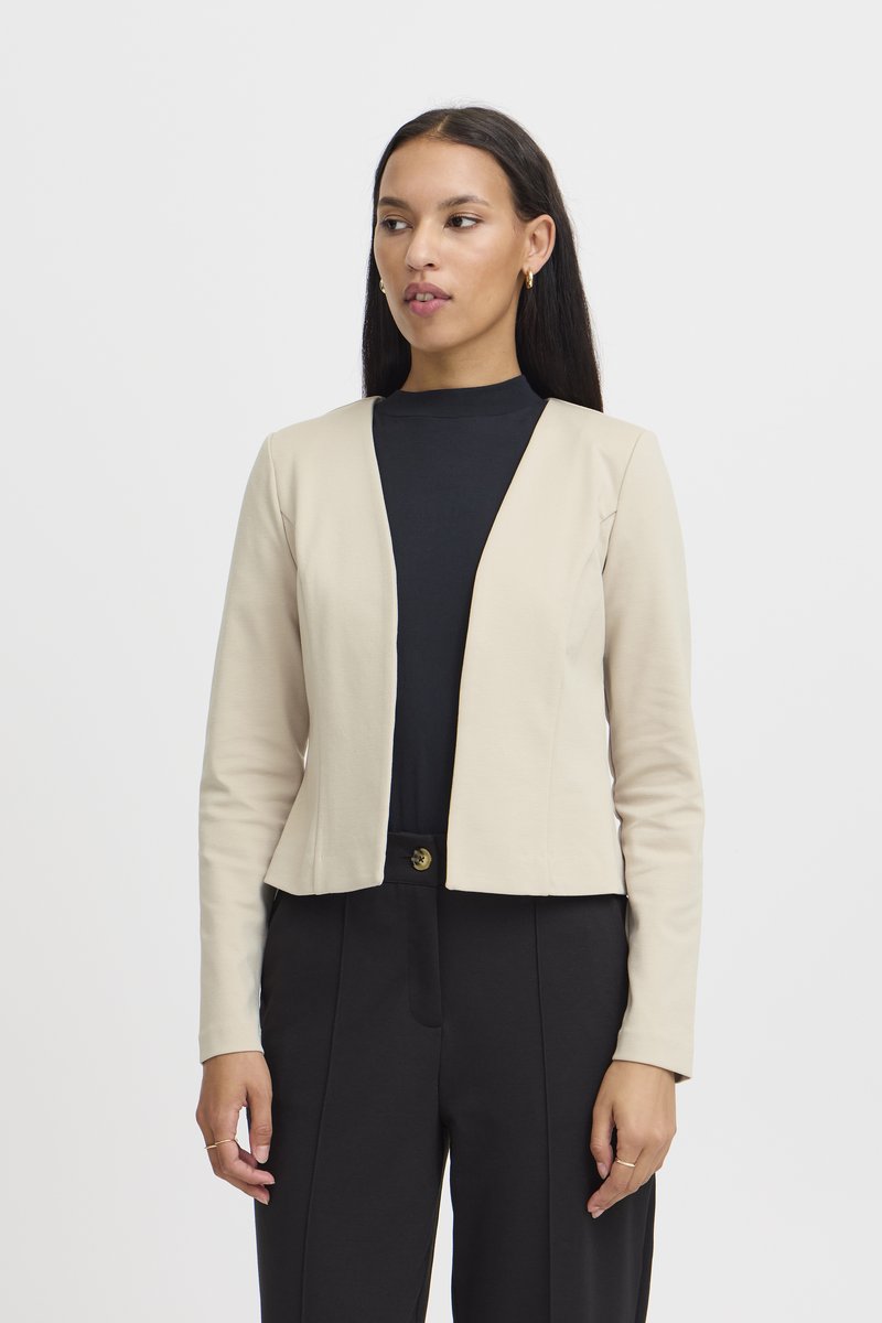 Mujer que lleva una blazer beige abierta sobre una blusa negra y pantalones negros, de pie frente a un fondo blanco liso.
