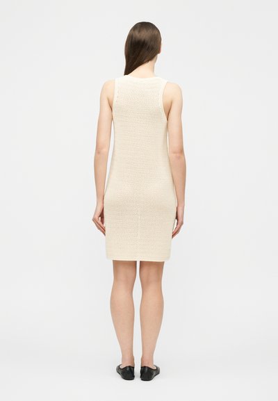 Γυναίκα που στέκεται με την πλάτη στραμμένη, φορώντας ένα αμάνικο, κρεμ πλεκτό φόρεμα μήκους μέχρι το γόνατο και μαύρα επίπεδα παπούτσια, μπροστά σε ένα απλό λευκό φόντο.