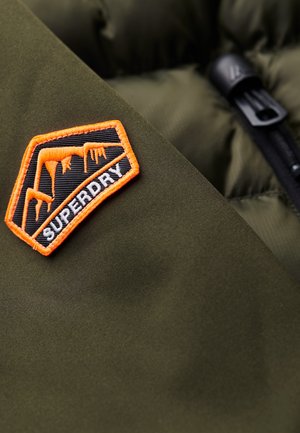 Prenda acolchada de color verde olivo oscuro con un parche del logo de Superdry en negro y naranja que presenta carámbano y un diseño geométrico. Cremallera visible.