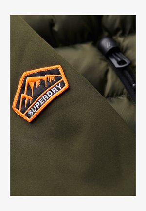 Prenda acolchada de color verde olivo oscuro con un parche del logo de Superdry en negro y naranja que presenta carámbano y un diseño geométrico. Cremallera visible.