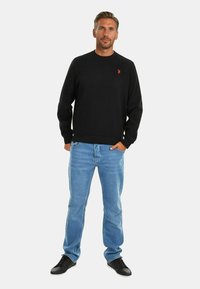 Black cotton sweatshirt with crew neck and red embroidered logo, paired with light blue denim jeans and black sneakers. Simple casual design.