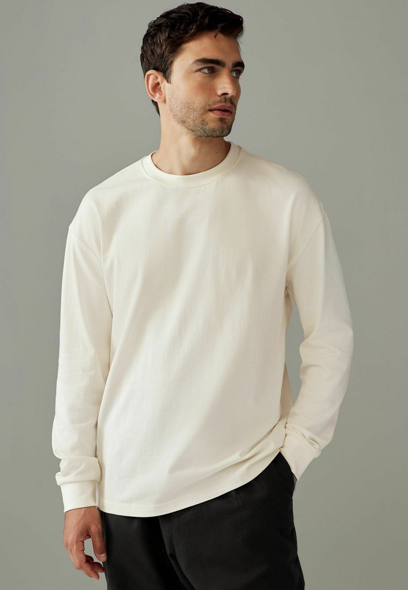 Cream long-sleeve t-shirt made of soft cotton, featuring a round neckline and relaxed fit, paired with black pants. Smooth texture.
