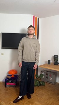 Beige zip-up hoodie with front pockets, embroidered text, paired with wide-leg dark denim jeans and shiny brown loafers.