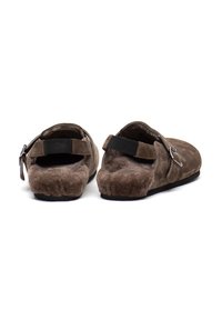 Zoccoli in suede marrone con superficie morbida e testurizzata, apertura sul retro, cinturino regolabile e suola in gomma, che mettono in evidenza un design minimalista.