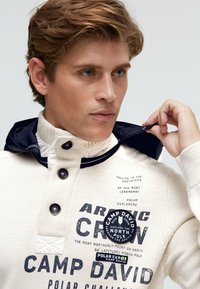 Weißer Strickpullover mit marineblauen Akzenten, ausgestattet mit einem hohen Kragen, drei Knöpfen und Aufnähern. Der Text umfasst Marken- und Explorationsthemen.