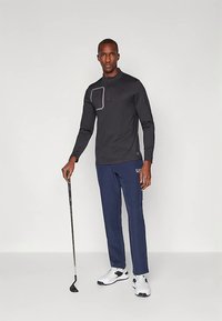 Juodas ilgomis rankovėmis marškiniai ir mėlynos kelnės vilkintis vyras laiko golfo lazdą, stovėdamas prieš paprastą šviesią foną.