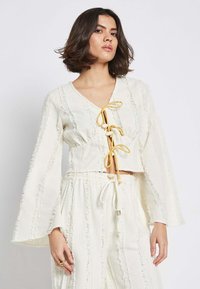 Valge lühikesed varrukad blouse, millel on pikad kellukesed, sügava V-kaeluse ja kollaste sidemete detailid. Tekstuuriga kangas, millel on kulunud aktsendid.