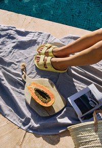 Person's legs in yellow sandals resting on a towel by a pool, with a halved papaya on a wooden board and an open magazine nearby.