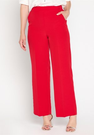 Kobieta nosząca jaskrawo czerwone spodnie o szerokich nogawkach, z ręką w kieszeni i srebrne sandały na otwartym palcu, stojąca na tle białego tła.