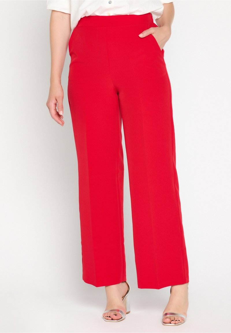 Femme portant un pantalon large rouge vif, main dans la poche et talons argentés à bout ouvert, debout devant un fond blanc.
