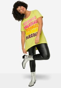 T-shirt giallo con testo ripetuto "PARADISE" in rosa e viola. Abbinato a leggings neri lucidi e stivaletti alla caviglia argentati.
