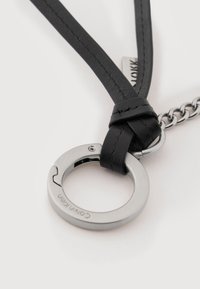 Ασημένιο κυκλικό μεταλλικό δαχτυλίδι με ανάγλυφη επωνυμία, προσαρτημένο σε μαύρο λουράκι από δέρμα και σύντομη ασημένια αλυσίδα σε λευκό φόντο.