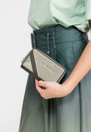 Persona sosteniendo una cartera con cremallera Anekke beige y gris, vistiendo pantalones de tiro alto verde oscuro y una blusa verde clara.