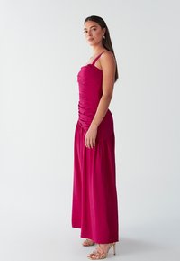 Vestido maxi fucsia confeccionado en tela suave con detalles fruncidos en el corpiño, tirantes finos y falda fluida. Combinado con sandalias metálicas de tiras.
