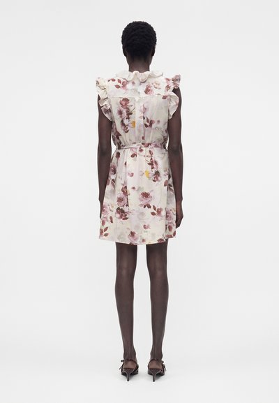 Γυναίκα που φοράει αμάνικο, flor αμάνικο φόρεμα με ζώνη, με βολάν στους ώμους και γιακά, στέκεται γυρισμένη με την πλάτη με καφέ τακούνια.