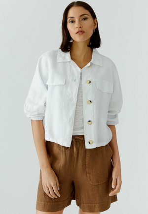 Femme portant une veste-chemise blanche boutonnée sur un haut blanc et un short marron, debout devant un fond clair uni.