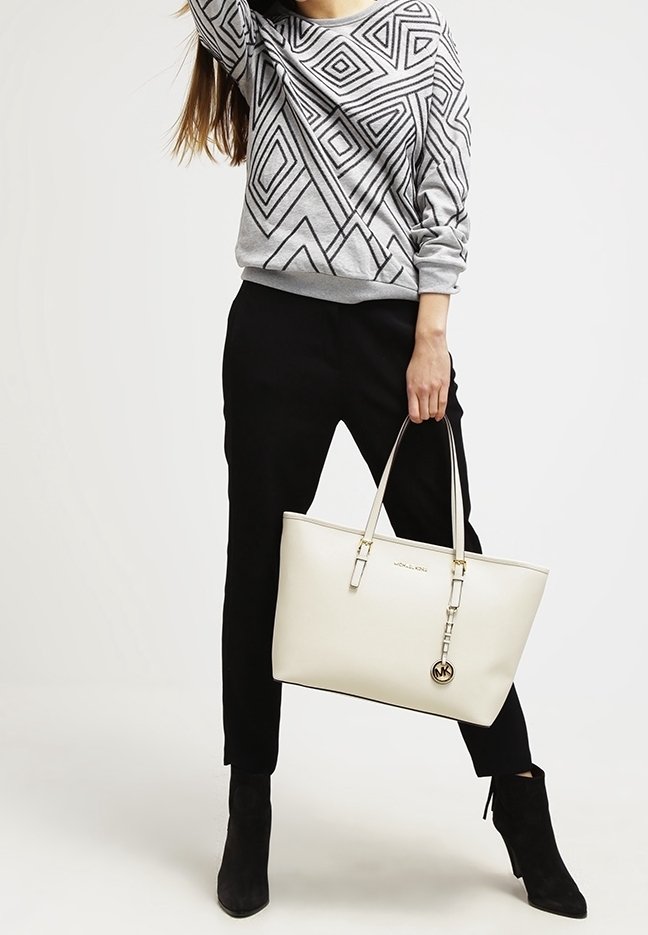 Sac fourre-tout en cuir blanc avec quincaillerie dorée, mettant en avant un accent de porte-clés. Le modèle porte un pull géométrique gris et un pantalon noir fuselé.