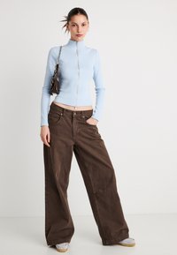 Ανοιχτό μπλε πλεκτό τοπ με ριμπ ύφανση, ψηλό γιακά και φερμουάρ μπροστά, συνδυασμένο με καφέ άνετο παντελόνι. Αξεσουάρ περιλαμβάνουν μια μικρή τσάντα χειρός.