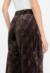 Pantaloni in velluto marrone con elastico in vita, caratterizzati da un motivo logo ripetuto e dettagli con tasche laterali. Texture liscia e vestibilità rilassata.