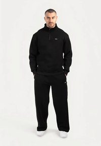 Felpa nera in pile con cerniera e colletto alto, abbinata a pantaloni della tuta neri coordinati. Logo bianco sul petto e sui pantaloni. Sneakers bianche.