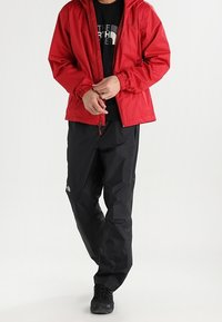 Czerwona wodoodporna kurtka z kapturem, z zamkiem błyskawicznym, ściągnięty dół. Czarne wodoodporne spodnie z akcentem w postaci logo, w zestawieniu z czarnym obuwiem.