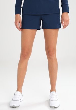 Mujer con pantalones cortos deportivos azul marino y una camiseta de manga larga a juego, con zapatillas blancas, de pie sobre un fondo blanco liso.