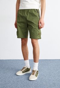 Green shorts with an elastic waistband, featuring side pockets, a zipper detail, and a logo accent, paired with beige and green sneakers.