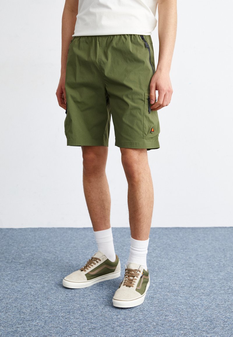 Green shorts with an elastic waistband, featuring side pockets, a zipper detail, and a logo accent, paired with beige and green sneakers.
