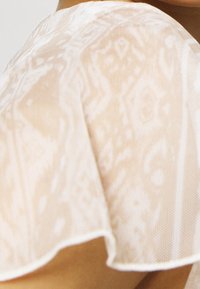 Tissu léger et transparent de couleur beige pâle avec une texture subtilement imprimée. Présente un ourlet délicatement froncé et une apparence légère et délicate.