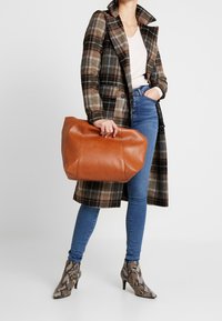 Brązowa skórzana torba typu tote o strukturalnym kształcie. Posiada gładką teksturę i rączkę u góry, uzupełnioną płaszczem w kratę i niebieskimi dżinsami.