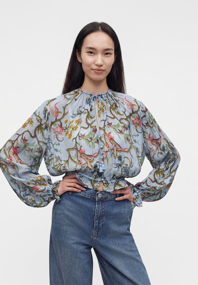 Femme aux longs cheveux foncés porte un chemisier à manches longues à motif floral pastel et un jean bleu, debout les mains sur les hanches devant un fond uni.