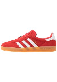 Röd mockasinsneaker med vita läderstripes; texturerad tunga, orange gummisula; har "Gazelle"-märkning och Adidas-logotyp.