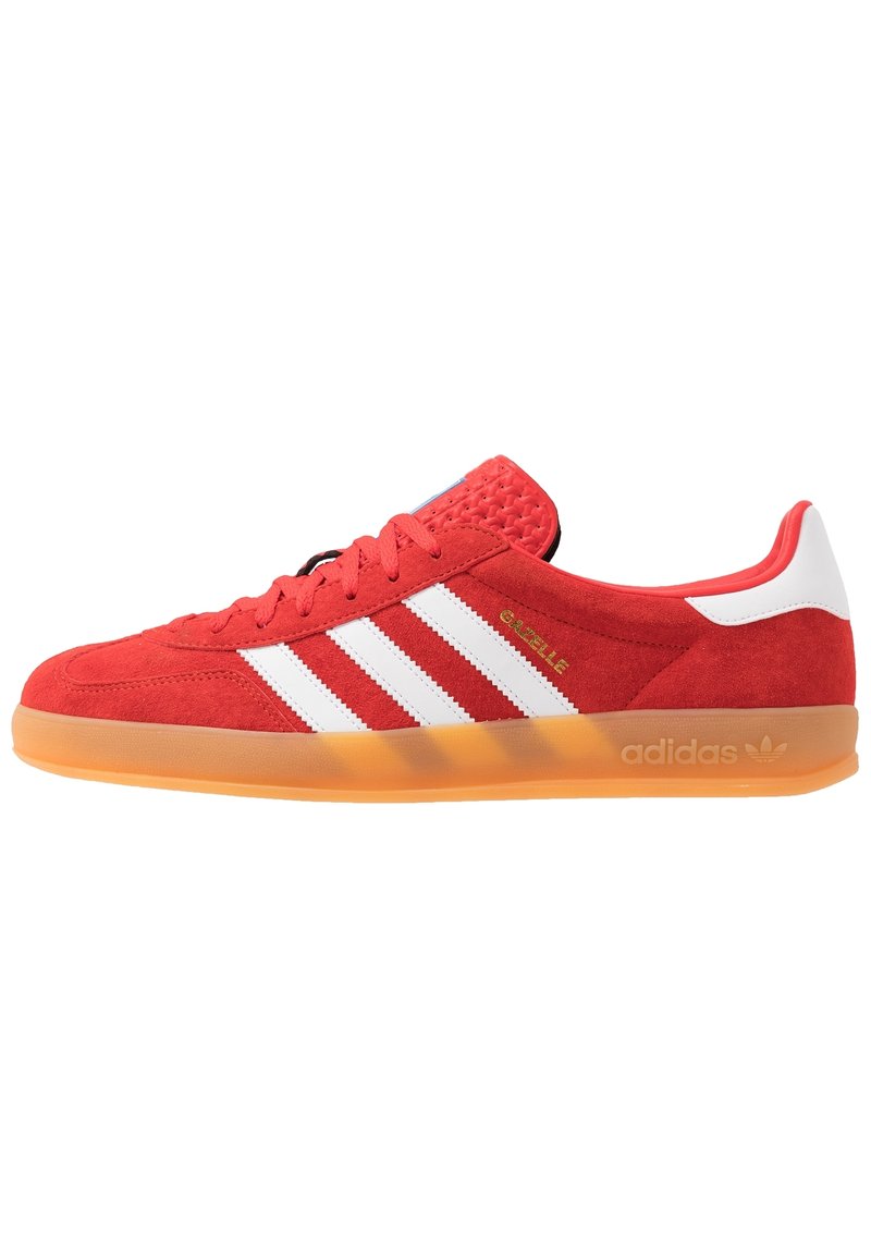 Röd mockasinsneaker med vita läderstripes; texturerad tunga, orange gummisula; har "Gazelle"-märkning och Adidas-logotyp.