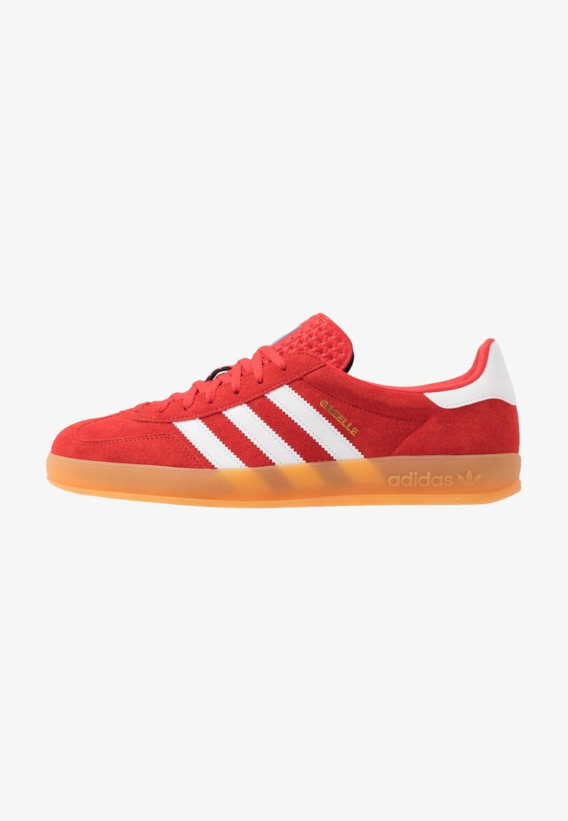 Röd mockasinsneaker med vita läderstripes; texturerad tunga, orange gummisula; har "Gazelle"-märkning och Adidas-logotyp.