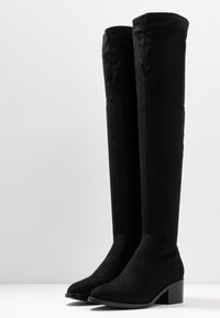 Botas de tela negra hasta la rodilla con textura suave, punta afilada y tacón cuadrado; costuras mínimas y sin adornos visibles.