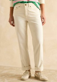 Kreemivärvi sirged püksid kõrge vöökohaga, millel on ees münditasku ja rullitud varrukaotsad, koos beežide tossudega.