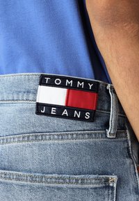 Šviesiai mėlyni džinsai su juodu ir raudonu užtrauktuku, kuriame yra "TOMMY JEANS" tekstas; matomas siuvimas ir užpakalio kišenės detalė.