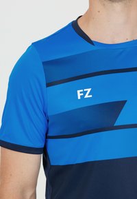 Mies, der ein blaues Sportshirt mit dunkelblauen geometrischen Mustern und weißen "FZ"-Buchstaben auf der Brust trägt.