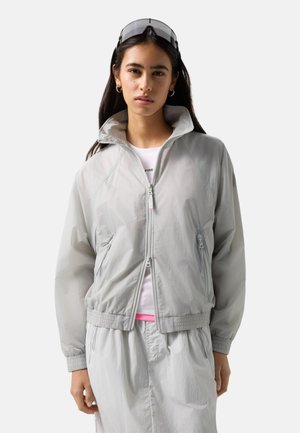 Young woman wearing light gray zip-up jacket and matching skirt, with sunglasses pushed up on her head, standing against a plain background.