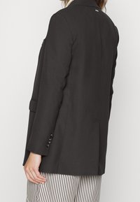 Musta räätälöity bleiseri, jossa on jämäkkä muotoilu, lovelliset kaulukset ja kolme nappiakselia hihoissa, käytetty raidoitettujen housujen päällä.