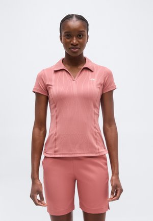 Junge Frau trägt ein rosa Polohemd mit kurzen Ärmeln und dezenten Wellenmustern sowie passende Shorts, steht vor einem einfarbigen Hintergrund.