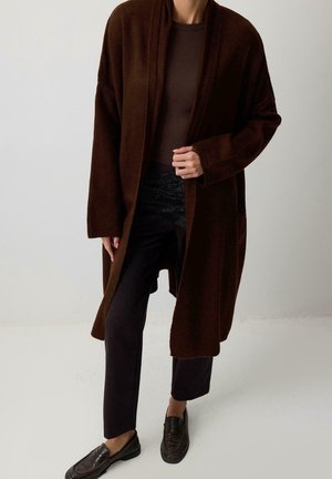 Personne portant un long cardigan ouvert marron foncé, un pantalon noir et des mocassins marron foncé, debout devant un fond clair uni.