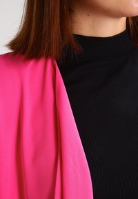 Fuchsia-draperad plagg över en svart ribbad topp. Tyget verkar slätt, med en flödande design och hög halsringning, vilket fokuserar på kontrast i färg.