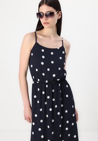Rochie fără mâneci, de culoare albastru închis, cu buline albe, având talie ajustată și fustă fluidă. Modelul poartă ochelari de soare închiși la culoare.