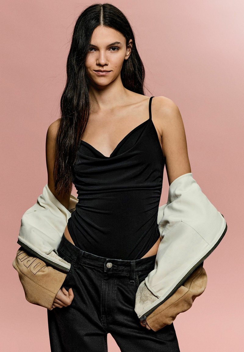 Body negru cu gât drapat, asortat cu o jachetă oversized albă și marron. Modelul poartă blugi negri cu talie înaltă pe un fundal roz.