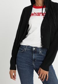 Černý pletený kardigán s ribkovanou texturou, s velkými knoflíky; nošený přes bílý tričko s červenými akcenty a modré džíny.