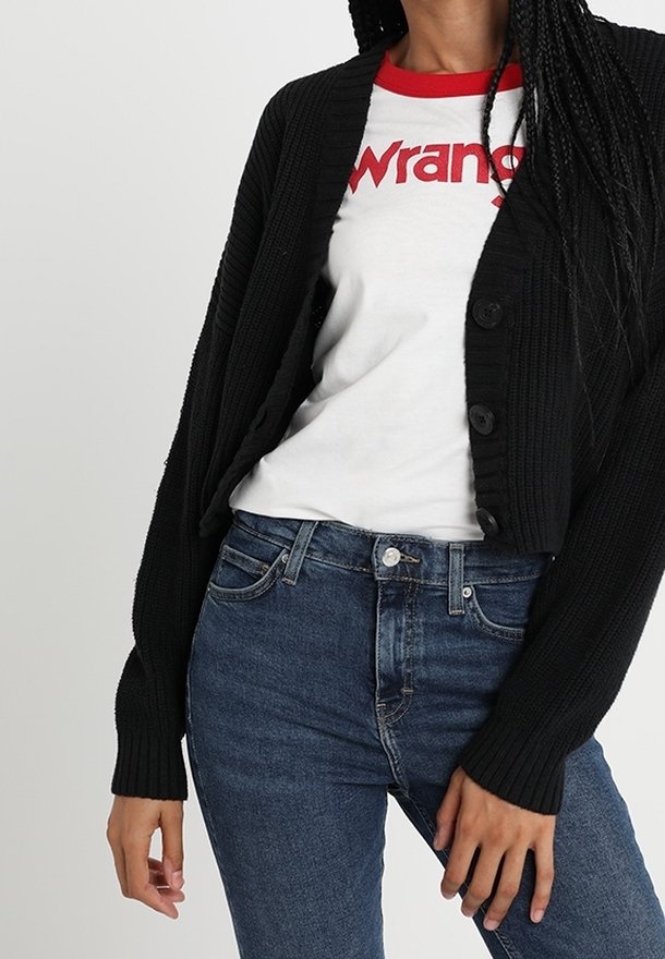 Černý pletený kardigán s ribkovanou texturou, s velkými knoflíky; nošený přes bílý tričko s červenými akcenty a modré džíny.