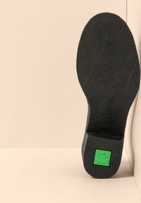 Suela de zapato negra con un logo de rana verde incrustado cerca del talón, posicionada de pie contra un fondo beige.